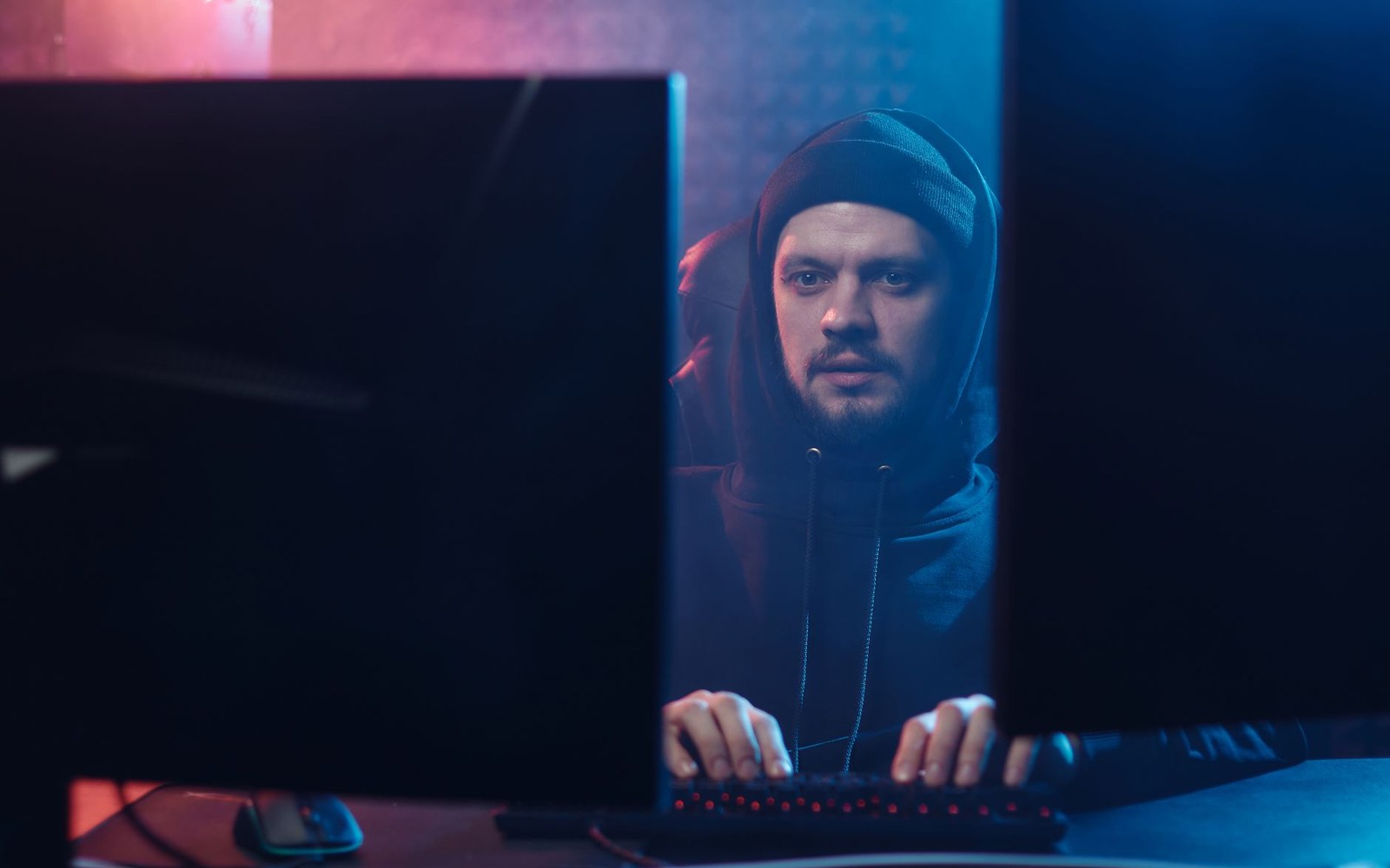 An adult developer's hands on a mechanical keyboard at night, the face partially lit by the blue-white glow of a large monitor showing source code