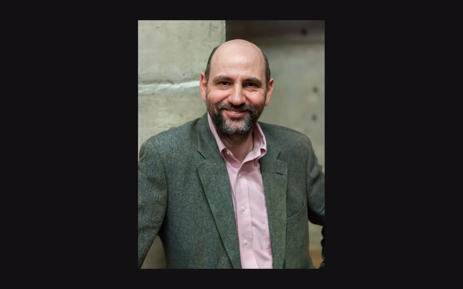 Bearded man in a tweed blazer and pink shirt smiling against a concrete wall