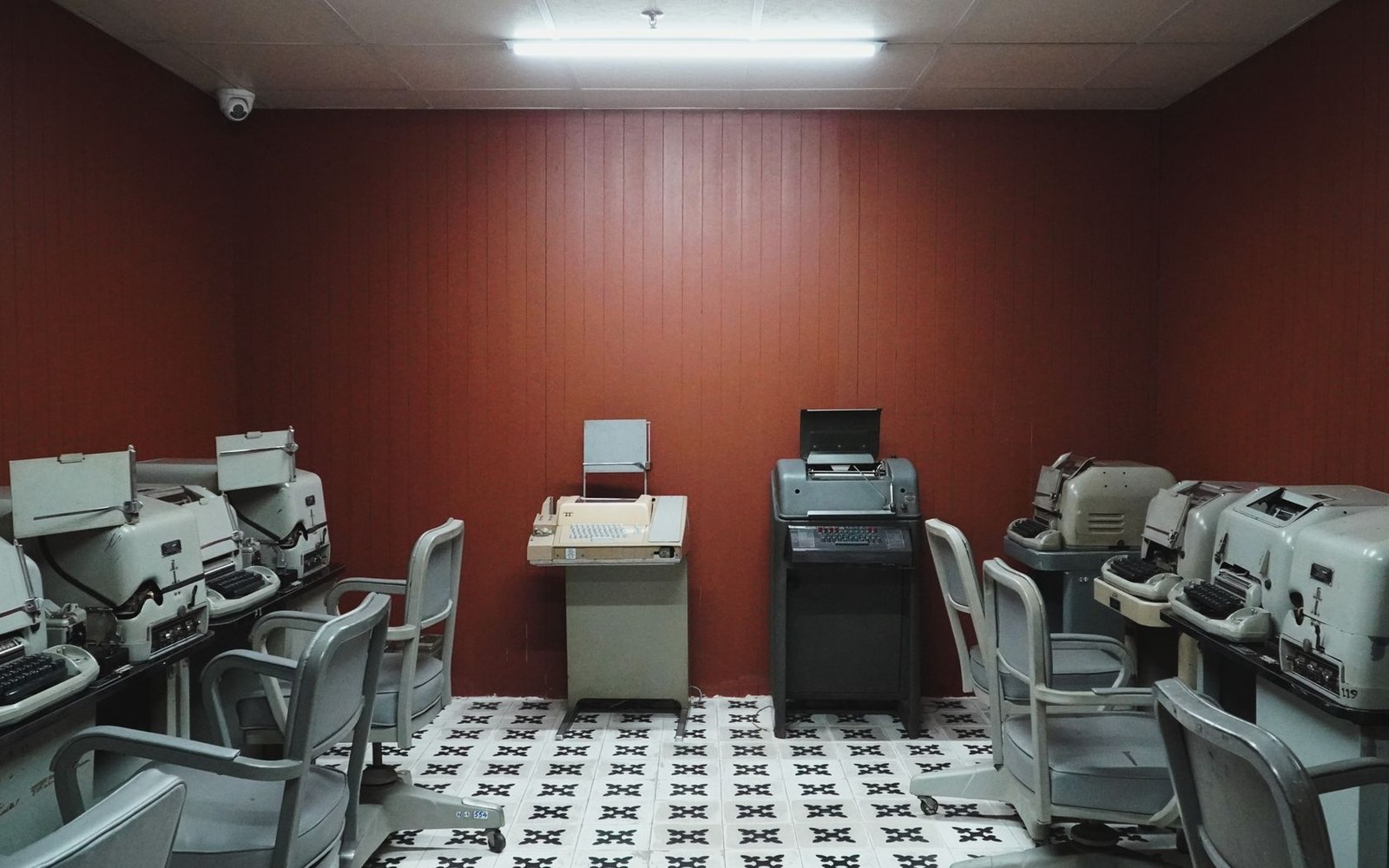 Row of vintage teletype machines and chairs lining a wood-paneled room with patterned floor