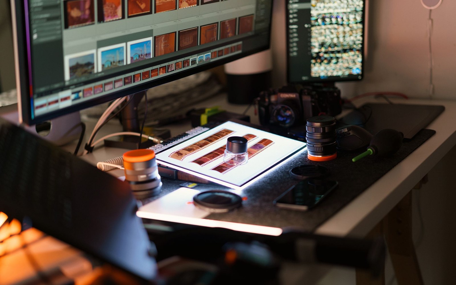 Film negatives on a light box beside camera lenses and photo-editing monitors on a desk