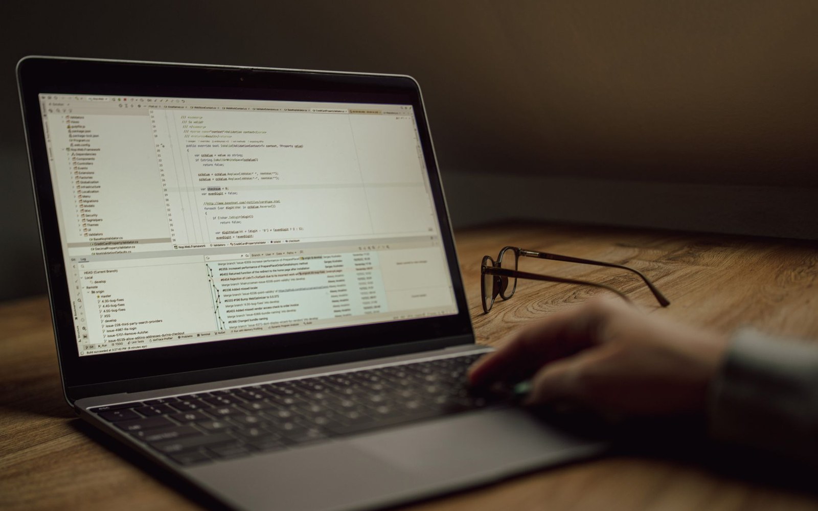 A hand rests on a laptop trackpad beside glasses, with code displayed on screen